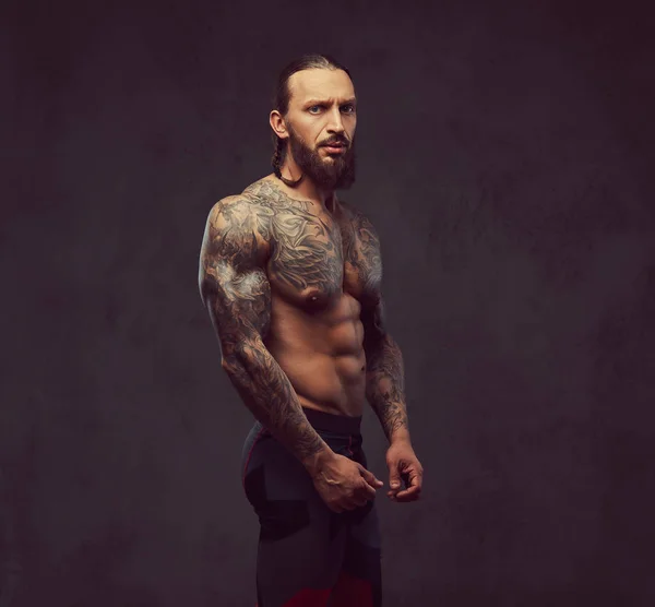 スポーツ ズボンでスタイリッシュな散髪と筋肉裸髭刺青男性の肖像画 暗い背景に分離 — ストック写真