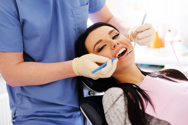 Dentist in blue uniform and white gloves cares on a sexy brunette caucasian woman, stand behind her, look in her mouth with a little mirror.