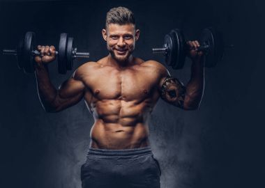 Dumbbells ile egzersizleri yapıyor gülümseyen güçlü şık vücut geliştirmeci, kolundaki dövme ile. Karanlık bir arka plan üzerinde izole.