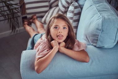 Uzun kahverengi saçlı ve delici göz atmak, mutlu küçük bir kız portresi evde kanepede yalan makinesinde dilini gösterir.