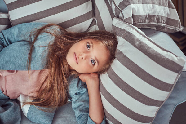 Portrait of a cute little girl with long brown hair and piercing glance, looking at a camera, lying on a sofa at home alone.