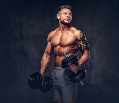 Dumbbells ile egzersizleri yapıyor güçlü şık vücut geliştirmeci, kolundaki dövme ile. Karanlık bir arka plan üzerinde izole.