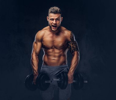 Dumbbells ile egzersizleri yapıyor komik şık vücut geliştirmeci, kolunda bir dövme ile. Karanlık bir arka plan üzerinde izole.