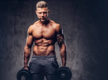 Kendinden emin bir bakışla kameraya bakarak bir stüdyoda dumbbells ile poz güçlü şık vücut geliştirmeci, kolundaki dövme ile. Karanlık bir arka plan üzerinde izole.