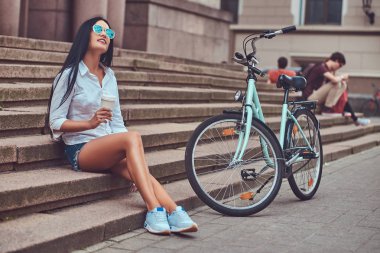 Bluz ve kot şort içinde güneş gözlüğü takıyor bir seksi esmer kadın üzerinde kahve keyfi bir bisiklet sürme sonra rahatlatıcı bir şehirde adımlar.