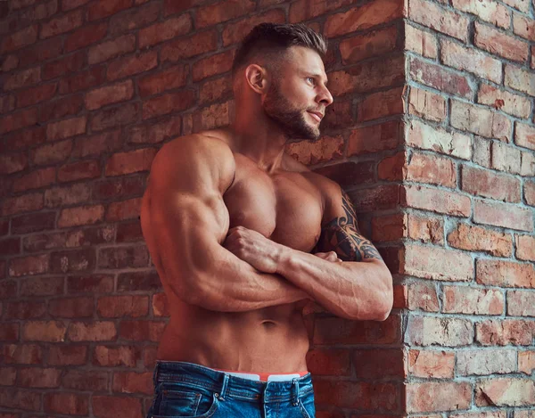 Portrait of a strong handsome shirtless male with muscular body, wearing jeans, standing with crossed arms, leaning against a brick wall in a room with a loft interior.