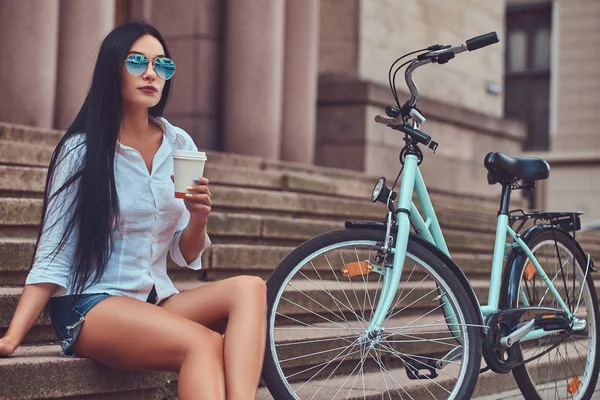 A sexy brunette female wearing blouse and denim shorts in sunglasses, relaxing after riding on a bicycle, drink coffee on steps in a city.