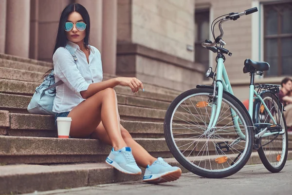 A sexy brunette female wearing blouse and denim shorts in sunglasses, relaxing after riding on a bicycle, sitting with a cup of coffee on steps in a city.