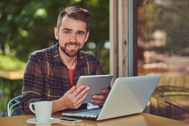Bir dizüstü bilgisayar üzerinde çalışan ve bir tablet bir kafeterya içinde tutarak Polar gömlek giymiş bir gülümseyen yakışıklı şık erkek çevirmen şık saç ve sakal,.