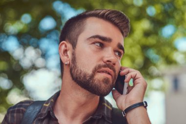 Yakın çekim portre bir genç güzel erkek Polar gömlek giymiş şık saç ve sakal, yanında bir smartphone şehir Park konuşuyor.