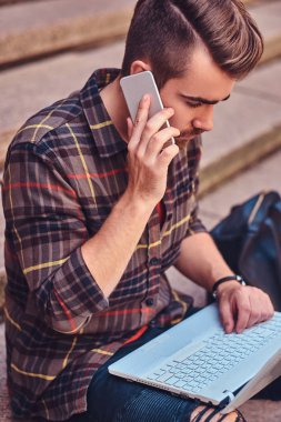 Bir Polar gömlek ve pantolon giyen güzel bir çevirmen ile şık saç ve sakal, bir dizüstü bilgisayar üzerinde çalışan ve basamaklarında oturan bir smartphone, konuşmak.