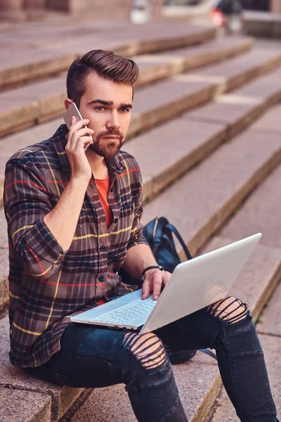 A beautiful freelancer with stylish haircut and beard, wearing a fleece shirt and jeans, working on a laptop computer and talk by a smartphone, sitting on steps.