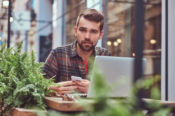 Portrait of a young freelancer with stylish haircut and beard, dressed in a fleece shirt, using a smartphone while working on a laptop computer, sitting in a cafe outdoors.