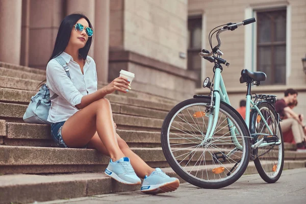 A sexy brunette female wearing blouse and denim shorts in sunglasses, relaxing after riding on a bicycle, sitting with a cup of coffee on steps in a city.