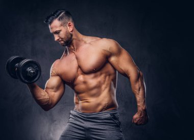 Uzun şık gömleksiz vücut geliştirmeci pazı ile bir stüdyoda ayakta bir dumbbell egzersiz yapıyor spor şort giymiş. Karanlık bir arka plan üzerinde izole.