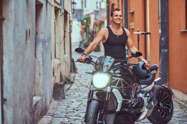 Siyah t-shirt ve jeans, dar bir eski Europe sokakta bir motosiklet yakın duran sevimli bir gülümseme ile yakışıklı bir vahşi motorcu giymiş.