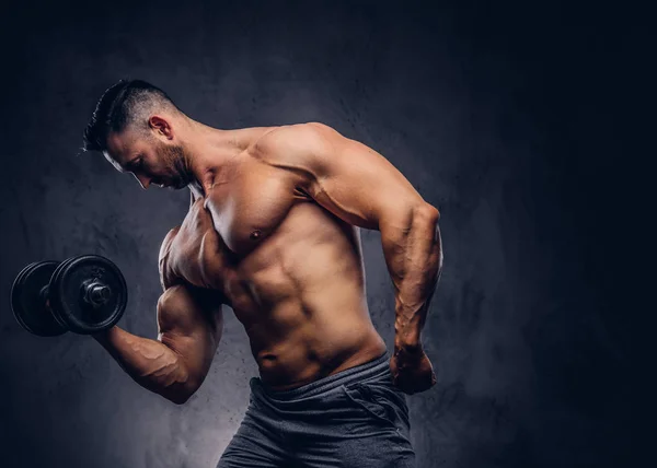 Tall stylish shirtless bodybuilder dressed in sports shorts, doing exercise on a bicep with a barbell, standing in a studio. Isolated on a dark background.