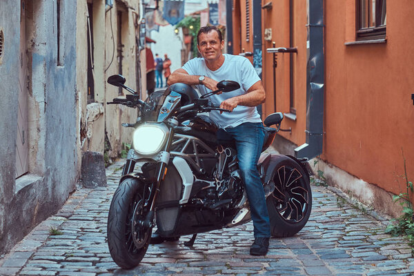 A handsome brutal biker dressed in a white t-shirt and jeans, with a charming smile, sitting on a motorcycle, in a narrow old Europe street.