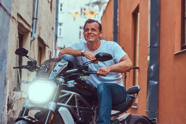 A handsome brutal biker dressed in a white t-shirt and jeans, with a charming smile, sitting on a motorcycle, in a narrow old Europe street.