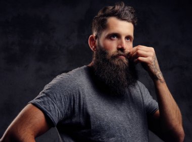 Karanlık bir arka plan üzerinde bir stüdyoda bir düşünce ile tam sakal ve gri bir t-shirt giymiş şık saç kesimi, dövmeli bir hipster portresi standları.