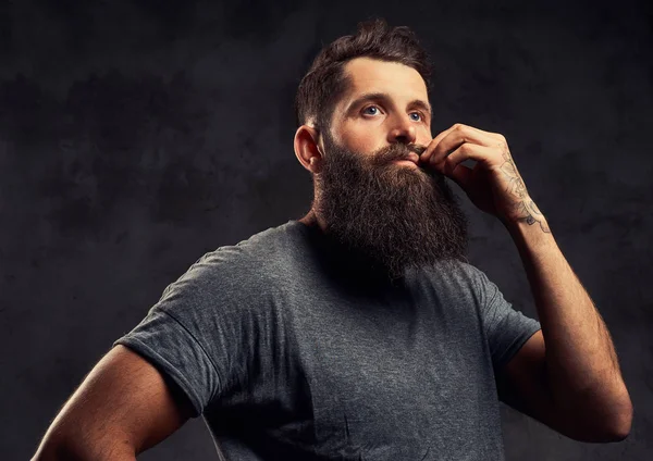 Portrait of a tattooed hipster with full beard and stylish haircut, dressed in a gray t-shirt, stands with a thinking look in a studio on a dark background.
