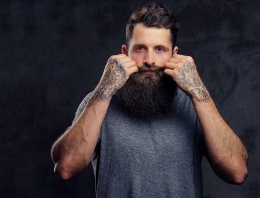 Karanlık bir arka plan üzerinde bir stüdyoda tam sakal ve kameraya, seyir gri tişört giymiş şık saç kesimi, dövmeli bir hipster portresi standları.