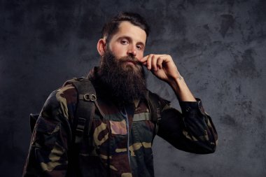 Yakışıklı bir gezgin, bir stüdyoda ayakta dövmeli bir silah ile trendy bir Kamuflaj ceketi giymiş sakallı. Karanlık bir arka plan üzerinde izole.