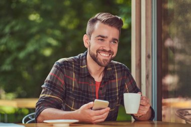 Yakışıklı bir moda erkek kahve içme ve kafede bir akıllı telefon tutarak Polar gömlek giymiş şık saç ve sakal,.