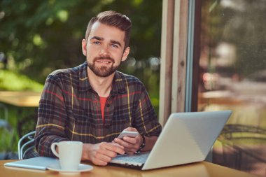 Bir dizüstü bilgisayar üzerinde çalışan ve bir kafeterya içine bir akıllı telefon tutarak Polar gömlek giymiş bir gülümseyen yakışıklı şık erkek çevirmen şık saç ve sakal,.