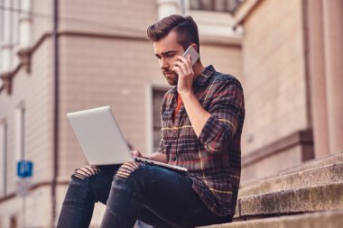 Bir Polar gömlek ve pantolon giyen güzel bir çevirmen ile şık saç ve sakal, bir dizüstü bilgisayar üzerinde çalışan ve eski bir bina karşı adımlar üzerinde oturan bir smartphone, konuşmak.