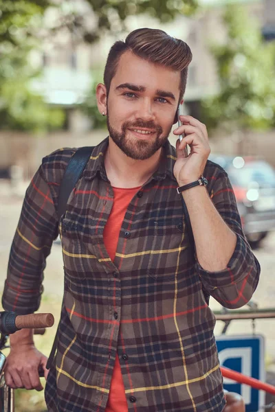 A handsome fashionable young male with stylish haircut and beard, wearing fleece shirt, stands in the parking lot for bicycles in the city park.