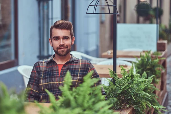Portrait of a smiling fashionable bearded male with a stylish haircut, dressed in a flannel shirt, sitting in a cafe outdoors.