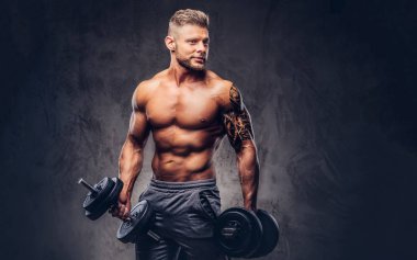 Dumbbells ile egzersizleri yapıyor güçlü şık vücut geliştirmeci, kolundaki dövme ile. Karanlık bir arka plan üzerinde izole.