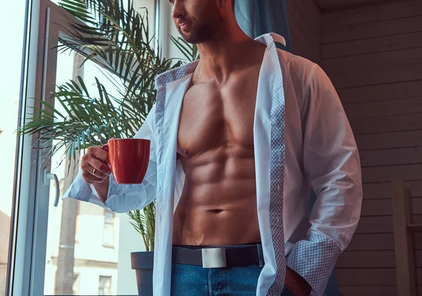 Unbuttoned シャツとジーンズに身を包んだ筋肉質のボディを持つ背の高いひげを生やした男性のクローズ アップの肖像画が窓の外見ている朝のコーヒーのカップを飲む — ストック写真
