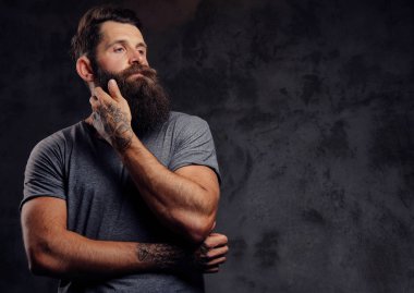 Karanlık bir arka plan üzerinde bir stüdyoda bir düşünce ile tam sakal ve şık saç kesimi, gri bir t-shirt giymiş bir hipster portresi standları.