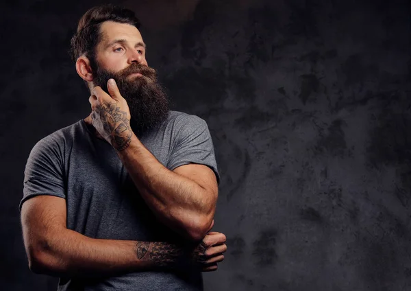 Portrait of a hipster with full beard and stylish haircut, dressed in a gray t-shirt, stands with a thinking look in a studio on a dark background. — Stock Photo, Image