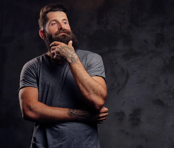 Retrato de un hipster con barba completa y corte de pelo elegante, vestido con una camiseta gris, se levanta con una mirada pensativa en un estudio sobre un fondo oscuro . — Foto de Stock