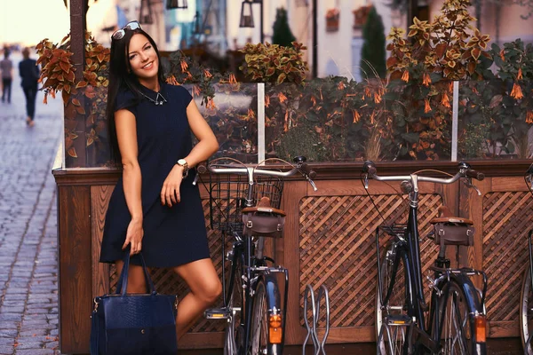 Усміхнена сексуальна брюнетка в стильній чорній сукні в сонцезахисних окулярах, тримає чорну сумку, позуючи біля велосипедів на фоні кафе . — стокове фото