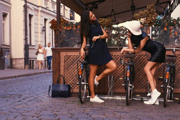 Две сексуальные брюнетки в стильных черных платьях, позируют возле велосипедов на фоне кафе . — стоковое фото