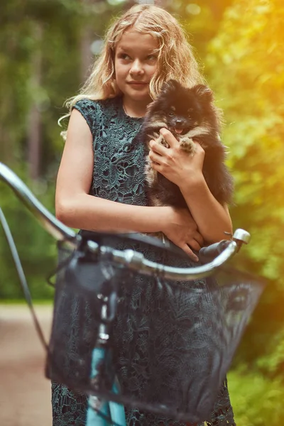 Retrato de una niña rubia en un vestido casual, sostiene lindo perro spitz, en un parque . — Foto de Stock