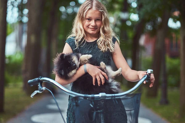 Porträt eines kleinen blonden Mädchens in einem lässigen Kleid, hält niedlichen Spitz-Hund. Fahrradtour im Park. — Stockfoto
