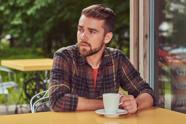Un hombre guapo de moda con corte de pelo elegante y barba, con camisa polar, beber café en la cafetería . — Foto de Stock