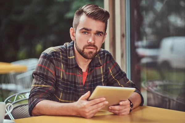 Un beau pigiste masculin à la mode avec une coiffure et une barbe élégantes, portant une chemise polaire, travaillant sur une tablette à l'intérieur d'un café . — Photo