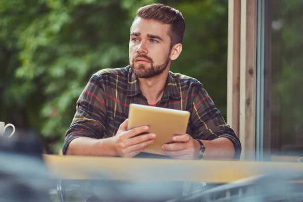 Um belo freelancer masculino elegante com corte de cabelo elegante e barba, vestindo camisa de lã, trabalhando em um computador tablet dentro de um café . — Fotografia de Stock