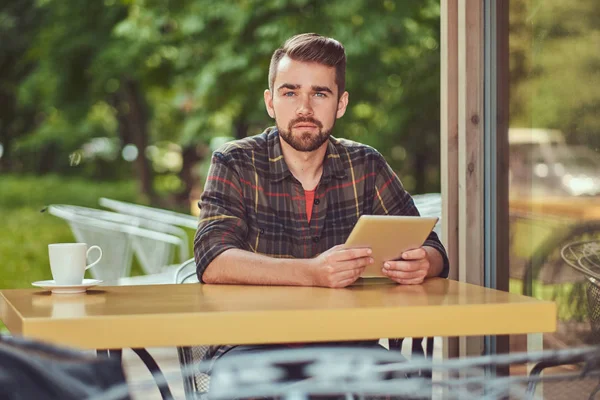 Um belo freelancer masculino elegante com corte de cabelo elegante e barba, vestindo camisa de lã, trabalhando em um computador tablet dentro de um café . — Fotografia de Stock
