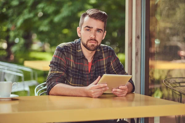 Un beau pigiste masculin à la mode avec une coiffure et une barbe élégantes, portant une chemise polaire, travaillant sur une tablette à l'intérieur d'un café . — Photo