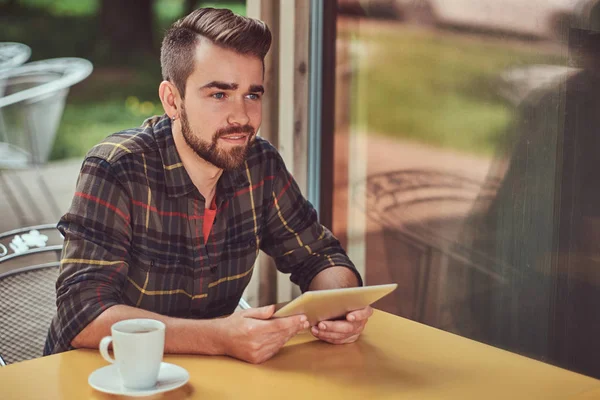 Un beau pigiste masculin à la mode avec une coiffure et une barbe élégantes, portant une chemise polaire, travaillant sur une tablette à l'intérieur d'un café . — Photo