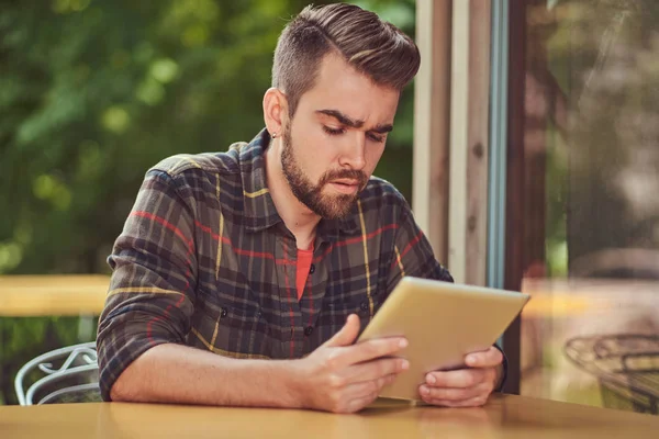 Um belo freelancer masculino elegante com corte de cabelo elegante e barba, vestindo camisa de lã, trabalhando em um computador tablet dentro de um café . — Fotografia de Stock