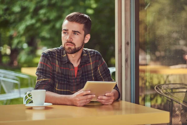 Um belo freelancer masculino elegante com corte de cabelo elegante e barba, vestindo camisa de lã, trabalhando em um computador tablet dentro de um café . — Fotografia de Stock
