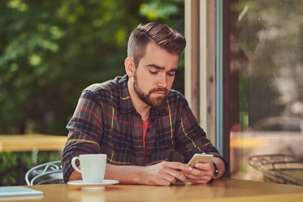 Un hombre guapo de moda con corte de pelo y barba con estilo, con camisa de lana, beber café y sostener un teléfono inteligente en la cafetería . —  Fotos de Stock